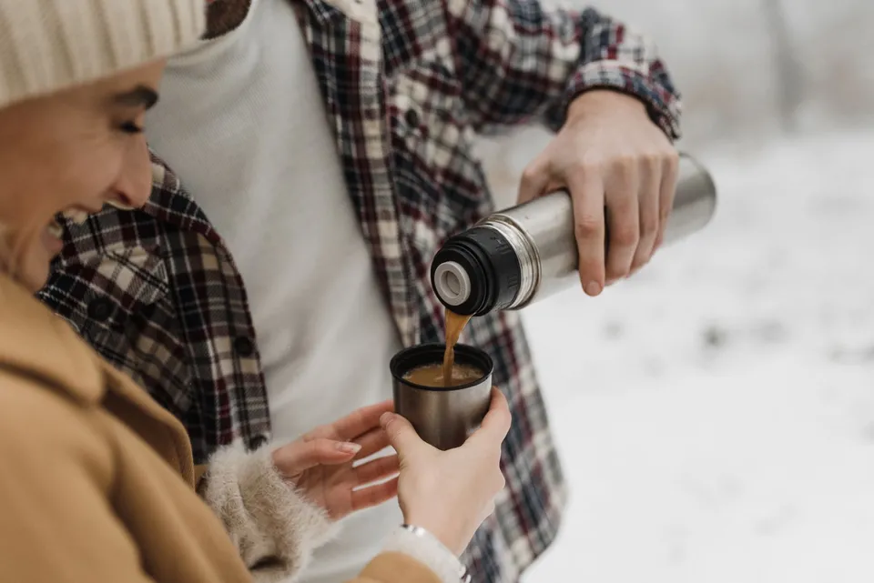 Two people pour coffee from a thermos in a snowy outdoor setting, enjoying a warm beverage.